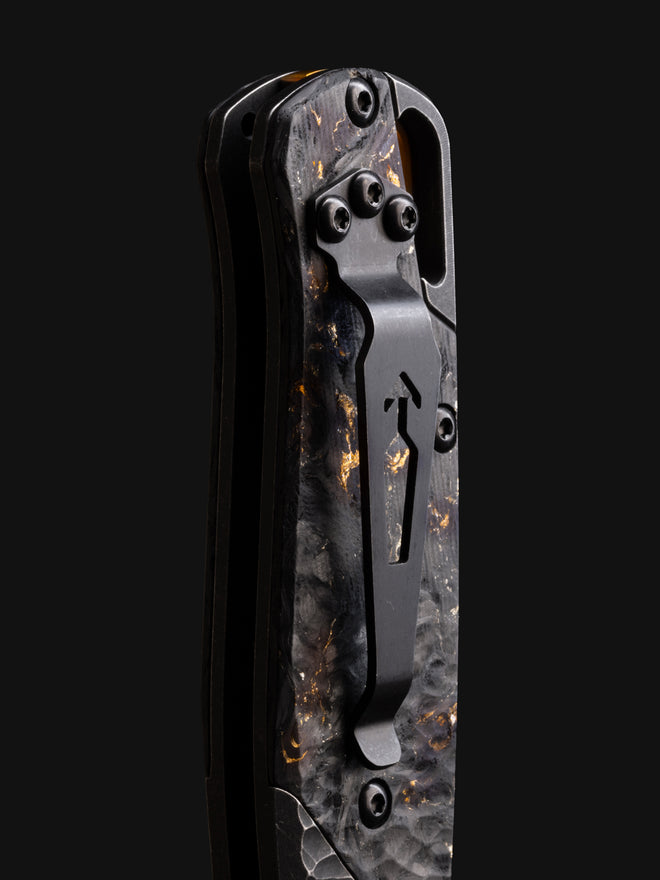 Close-up of a folding knife handle with black carbon fiber scales, gold flecks, and a matte black pocket clip on a dark background.