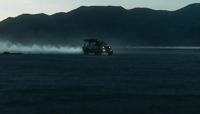White SUV with headlights on drives through dusty desert at dusk, kicking up dust against dark mountains.