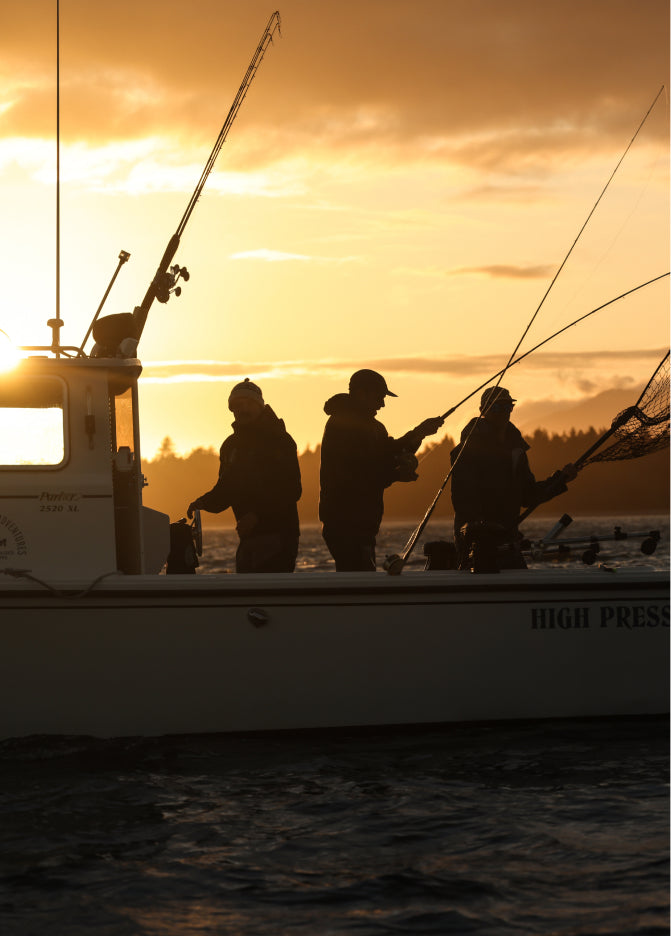 Three anglers on boat "High Press" at sunset, holding rods and net against orange sky.
