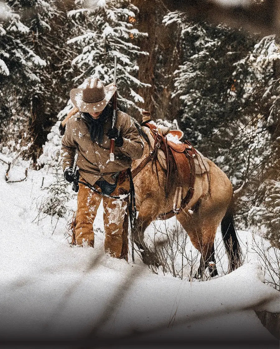 Cowboy in tan coat and hat holds rifle beside saddled palomino horse in snowy pine forest.